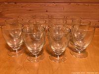 Set of twelve clear short footed water glasses arranged in two rows on a wooden surface, showing bowl shape and footed base.
