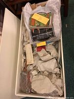 White rectangular toy box filled with toys and collectibles, including books, a yellow note, protective paper wrapping items