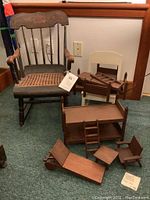 Two white kids chairs, two wooden child rockers, and multiple wooden doll furniture pieces placed on floor in front of wooden chair and wall.