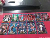 Image of 12 NBA basketball trading cards laid out on a red cloth surface with a laptop partially visible on the upper edge.