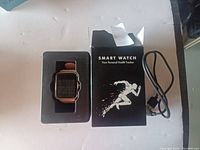 Smart watch in black tray with brown strap, rose gold case, black display, original black box with runner graphic and text, black USB charger cable.