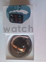 Top view of smart watch box with image of watch and actual smart watch with attached blue band, pink and black additional wrist bands, and charging cable packed inside box.