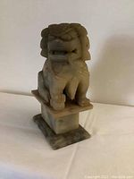 Front angled view of the carved stone Foo dog statue on pedestal, showing detailed face, mane, and pedestal base.