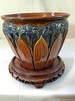 Front and side view of the ceramic planter showing blue and brown glaze with embossed floral or leaf designs and matching wooden stand with curved cutouts.