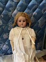 Front view of vintage porcelain doll seated against a patterned background showing white lace dress and braided hair.