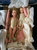 Two vintage dolls lying in a box with pink and striped fabric pillows and bedding, visible small shoes around the dolls.