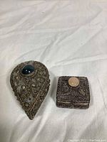 Two metal filigree decorative boxes, one teardrop-shaped with blue stone and one square with peach stone, shown closed.