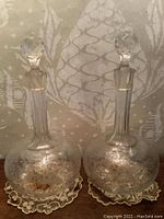 Pair of clear etched crystal decanters with floral and leaf patterns on wooden surface with lace doilies.