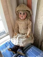 Full seated view of vintage porcelain doll wearing cream dress with lace and bonnet, shot near a window.