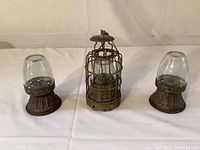 Front view of two copper base small oil lamps with clear hand-blown glass globes flanking one vintage wire cage oil lantern with glass globe and metal cover.
