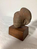 Front-right angled view showing the sculpted stone head with visible facial features and wood plinth beneath.