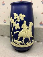 Side view of blue ceramic vase with cream raised relief of horseman and dog alongside a tree