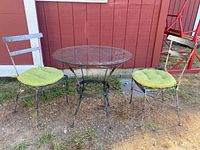 Full view of metal bistro set with glass top table and two green cushioned metal chairs, outside on dirt ground near red building.