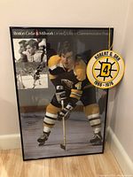 Full view of framed poster and attached fabric number plaque leaning against wall on hardwood floor.