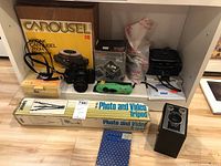 Photo showing the Kodak Carousel 760H slide projector in box, Sakar tripod in box, Ricoh KR-5 super II film camera, Canon PowerShot A480 digital camera, Nikon Coolpix S6100 digital camera, Polaroid I-Zone instant camera, slide viewer, camera case, and camera strap all arranged on floor and shelf.