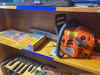 Photo of Husqvarna 240 chainsaw on wooden shelf with box containing spare chain and several hand tools underneath.