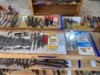 Overview of assorted drill bits, saw blades in packaging, pliers, screwdrivers, and miscellaneous hand tools on wooden table