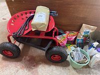Red garden seat with black wheels, rolled garden bird netting tied with blue ties on seat, two bags Vigoro Rose Food, beige bag Sea Magic, two plastic bottles, spray bottle, green bucket with gardening items, planter pot, and garden tools scattered around.