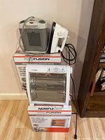 Image showing four Profusion infrared quartz radiant heaters stacked on top of their respective retail boxes along with one white electric clothing iron placed on the top heater. Heaters have metal mesh grills and control dials.