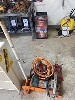 View of orange hydraulic floor jack on the ground with two red jack stands, coiled orange hose, and metal rods piled on top next to a white pole and shelving.