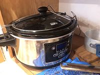Photo of Hamilton Beach stainless steel crockpot with glass lid on wooden surface with blue and metal utensils beside.