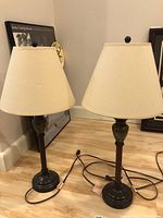 Two matching table lamps with beige shades and dark bases placed on wooden floor against wall.