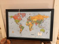 Framed world map with visible electrical cord hanging on a wall, showing full map surface and frame.
