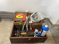 Wooden box filled with assorted plumbing supplies including flexible hoses, couplings, valves, solder, and tools.