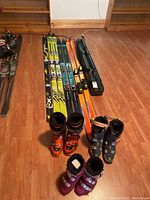 Photo shows seven pairs of skis laid out on floor with three pairs of boots in front, along with some ski carrying cases.
