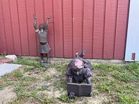 Wide shot showing both garden statues positioned outdoors near a building with red siding, girl statue lying with book and standing child statue with raised arms.