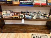 Wooden miniature village buildings of various styles on a wooden shelf with books behind them.
