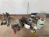 Wide view of all tiling and masonry tools arranged in a row on a concrete floor showing various hammers, chisels, spreaders, scrapers and buckets.