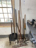 Vintage garden tools including shovels, a mallet, rakes, and other hand tools leaning against a wall near a window and workbench.