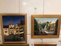 Two framed original oil paintings by Barbara Ripley on a white wall, the first depicts a village street scene, the second a waterside building and waterfall.
