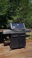 Full view of Weber Spirit propane gas grill showing black two-door cabinet, stainless steel side shelves, mounted on casters, outdoors on wooden deck