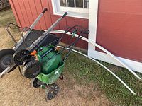 Metal wheelbarrow with two green seed spreaders and white PVC tubes leaning against red building wall.