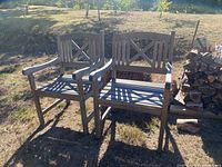Two weathered wooden outdoor chairs with armrests and cross-back design placed outdoors on grass beside stacked firewood.