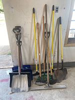 Full view of the yard and garden tools leaning against a white wall including shovels, rakes, a hoe, and a squeegee.
