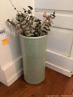 Full view of tall ceramic container holding dried flowers against a wall corner.