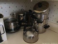 View of entire cookware set arranged on kitchen counter, showing multiple pots stacked, kettle in front, and frying pan to the side. Stainless steel with black handles and lids.