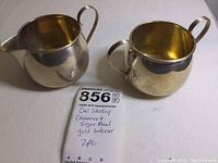 Top-front view of both pieces showing the creamer's spout and sugar bowl's open top with gold-toned interior, with a handwritten note confirming manufacturer and item details.