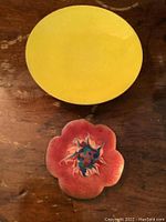 Photo of two enamel on metal plates placed on wood surface. Larger yellow round plate and smaller red flower-shaped plate with central floral pattern.
