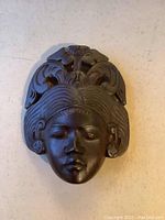 Front view of dark wood tribal face carving with detailed hair and headdress patterns