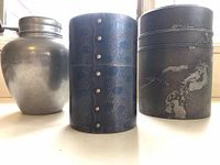 Photo showing three metal tea jars: one silver aluminum, one dark hammered metal with rivets, one taller dark embossed metal jar with scrolls and nature motifs.