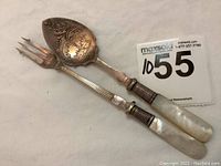 Two-piece mother of pearl handled pickle fork and ornate spoon on a neutral background with auction tag.