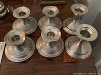 Photo showing six pewter candle holders arranged on wooden surface, displaying different styles and sizes
