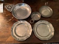 Top view of the five pewter items arranged on a wooden surface showing two scalloped edge plates, two round flat plates, and one porringer.