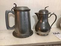 Front view of a pewter tankard with hinged lid and a covered pewter pitcher side by side on a shelf.