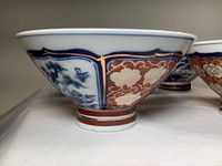 Side view of one rice bowl showing detailed traditional Asian design with blue and white scenic panels and reddish-orange floral panels with gold accents.