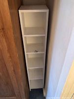 Tall, narrow white 5-shelf bookcase positioned between a wooden closet and a wall, showing its height and shelf structure.